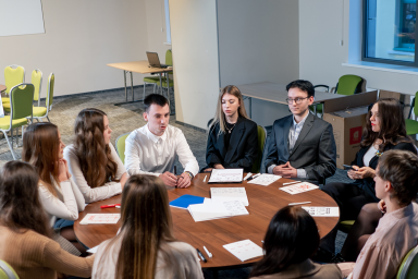 Photo of a group of senior students and adults having a discussion with pens and paper around a round table in a classroom