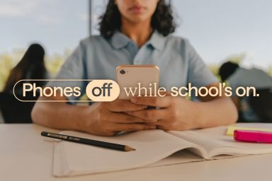 Student sitting at a desk, looking at a mobile phone. Text on screen 'Phones off while school's on.'