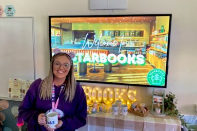 Teacher wearing a purple jumper and holding a coffee mug is standing in front of a screen with the text Welcome to Starbooks