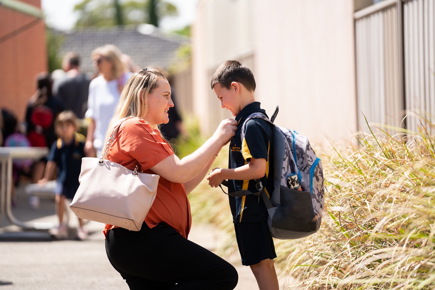 Mum kneeling down to speak to young boy in school uniform before he walks into school
