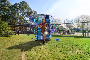 A smiling boy holding a football runs out of a giant smartphone displaying a video game, surrounded by colourful digital cubes, into a sunny school playground with green grass and trees.