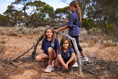 Photo of three students, one student is building a tree teepee, the others are sitting underneath the teepee