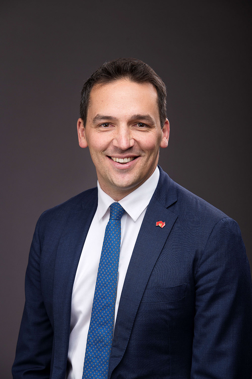 Blair Boyer, smiling and wearing a navy blue suit with a white shirt and blue tie.