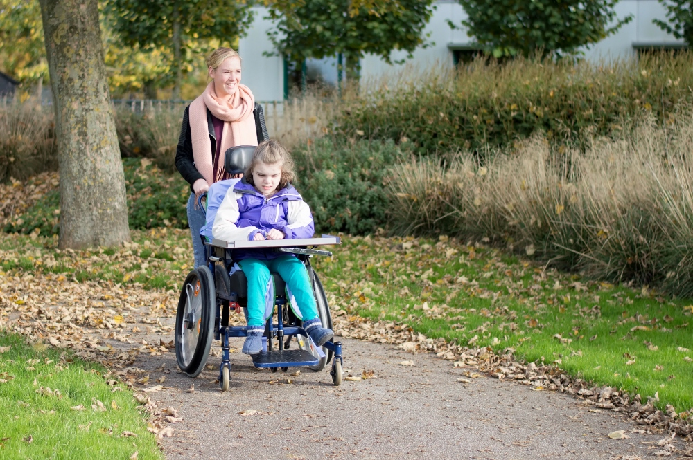 A child in a wheelchair, and a woman, go along a path in a park. Photo.