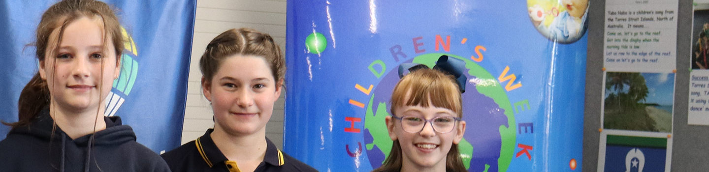 Three students with Minister for Education Awards, standing in front of a Children's Week banner.