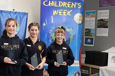 Three students with Minister for Education Awards, standing in front of a Children's Week banner.