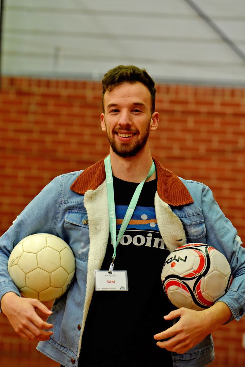 An adult holding 2 soccer balls in a gymnasium, smiling. 