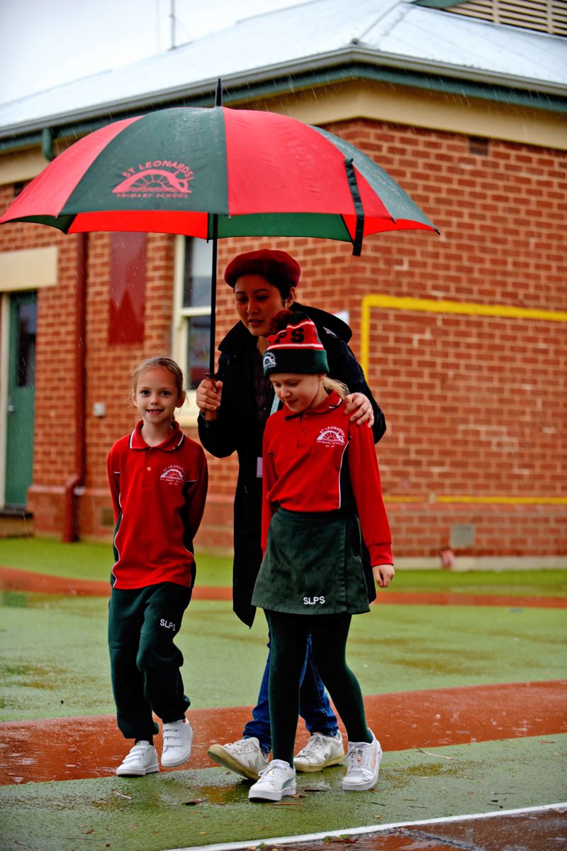 An adult volunteer with 2 students walking in the school yard with an umbrella.