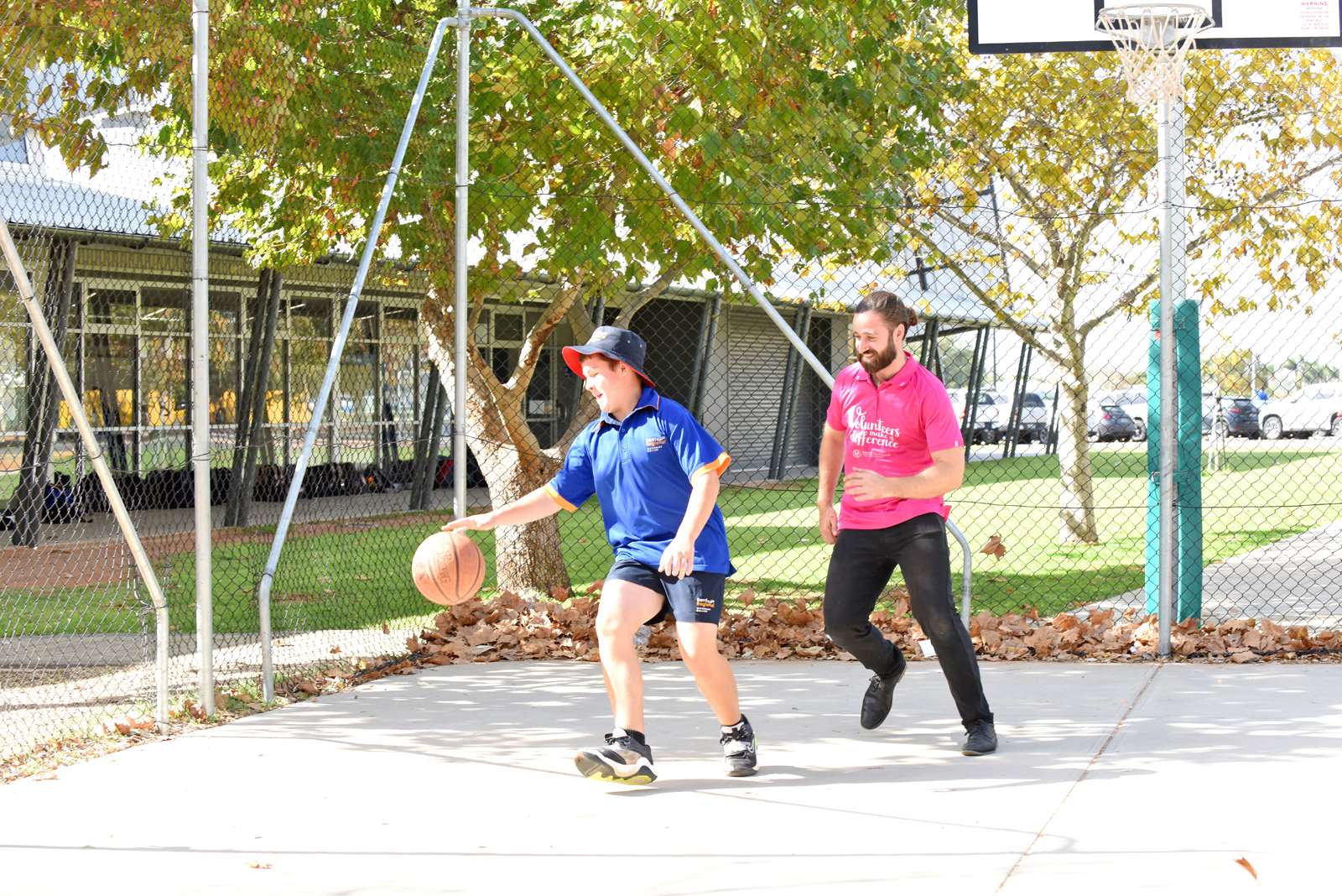 An adult volunteer playing basketball with a student on an outside court, it's a sunny day.