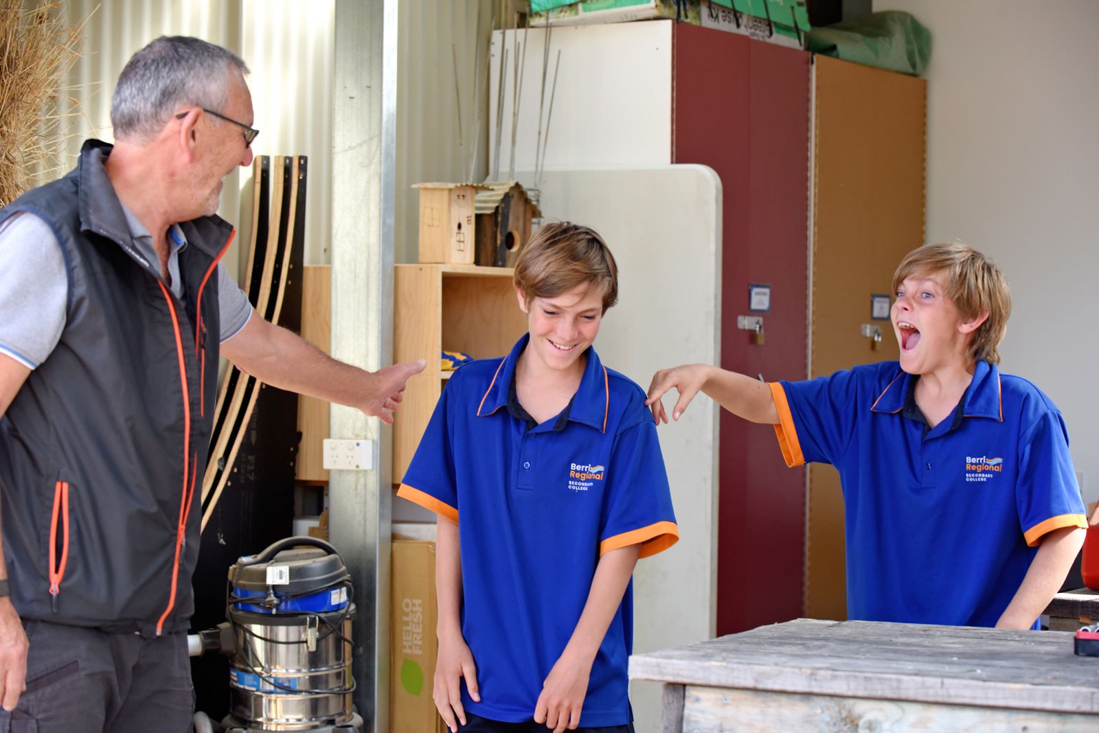 An older volunteer with 2 students in a wood workshop. They are laughing and smiling. 