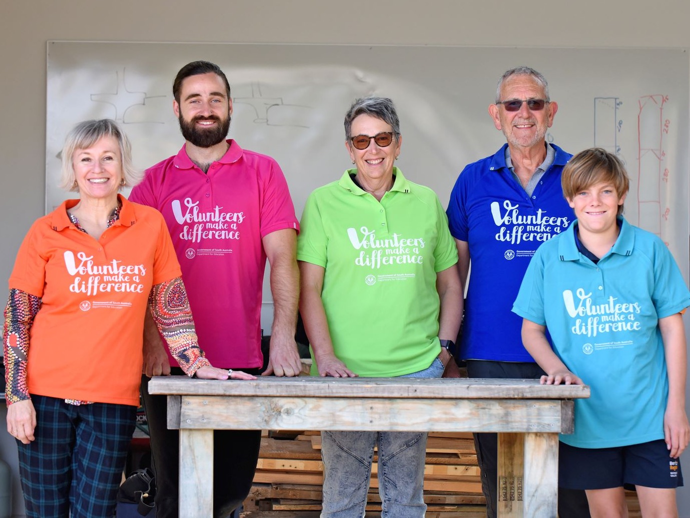 4 adults and a student volunteer wearing bright colour t-shirts, standing over a work bench smiling. 