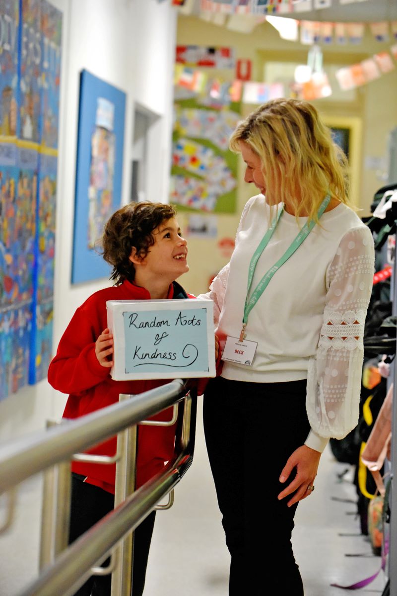 A young student is holding a sign that says 'random acts of kindness', looking up at an adult volunteer.