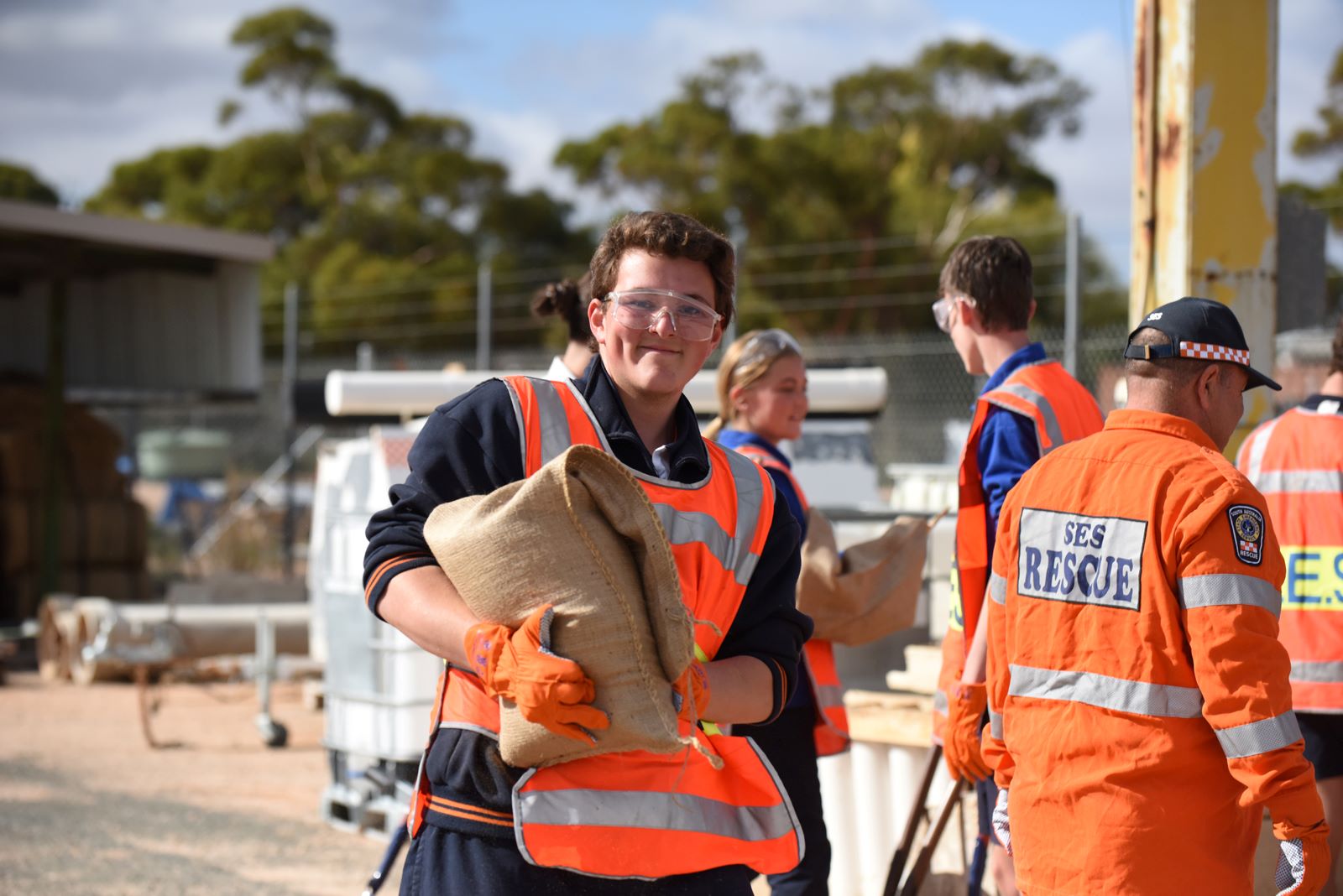 A student volunteer wearing safety glasses and high vis, carrying a sandbag. SES rescue crew members are behind them, they are smiling. 