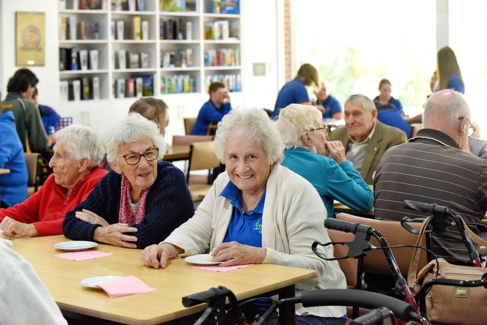 Elderly people in a group setting sitting at tables with their walkers next to them, and some students in the background.  