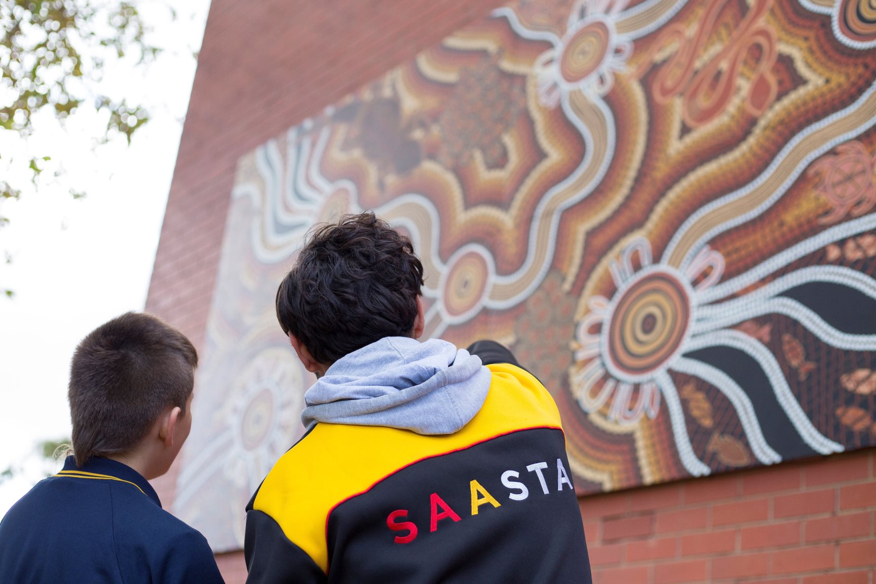 The backs of 2 people, looking and pointing at a large peice of Aboriginal art. The back of the jumper says SAASTA.