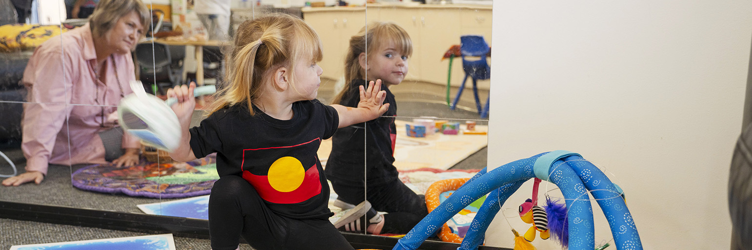 A child smiling at itself in the mirror while a parent smiles while watching